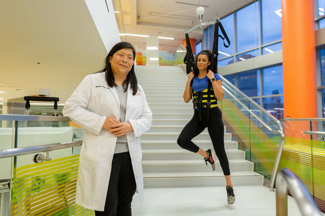 A woman of color in a lab coat in the foreground. A woman of color in a harness while trying out an experiment in the background.
