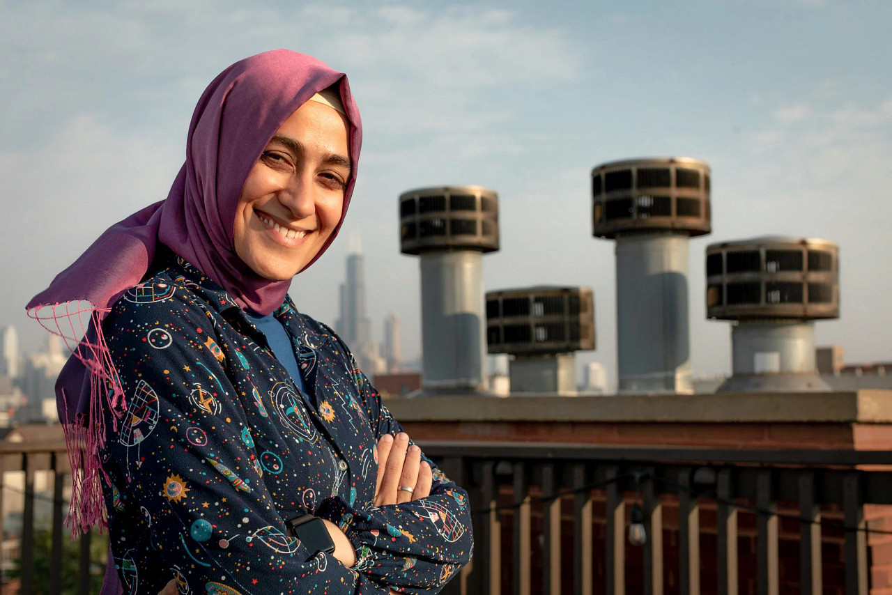 A woman in a head scarf smiling in front of architecture.